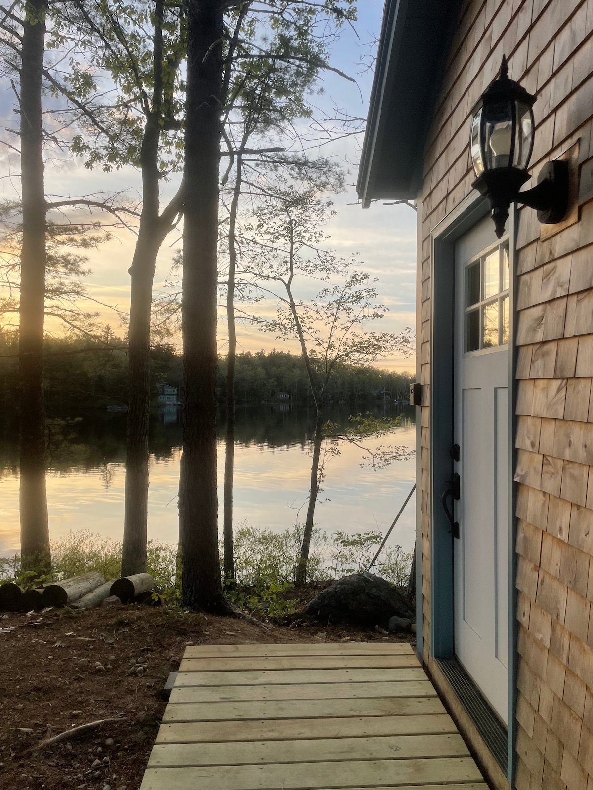 A wooden path leads to the entrance of a rustic cabin, framed by tall trees. The calm waters of Woods Pond reflect the soft colors of a sunset, creating a serene backdrop. The cabin’s door is accented by a wall-mounted lantern.