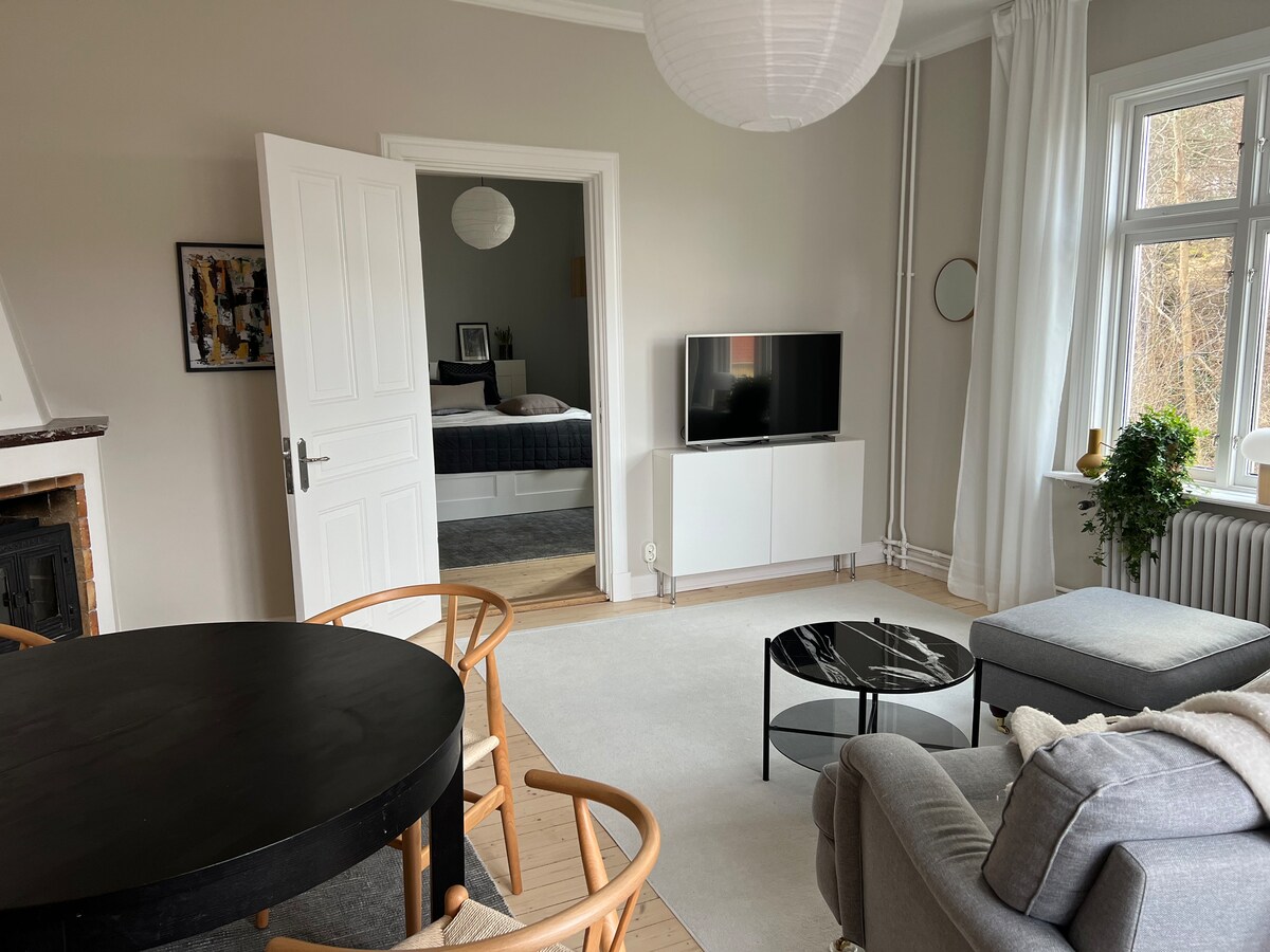 A spacious living area features a round dining table with six chairs, a comfortable grey couch, and a sleek black coffee table. Large windows allow natural light to fill the room. A doorway leads to a separate bedroom, enhancing the open layout.