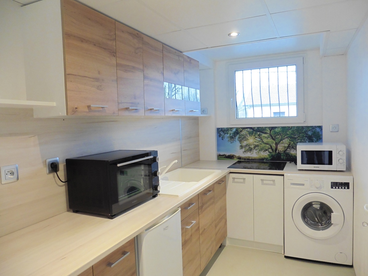 The kitchen is equipped with modern appliances, including a microwave and an oven. Light wood cabinetry complements the space, which features a window with a view of greenery. A washing machine is also present, along with a sink and countertop area.