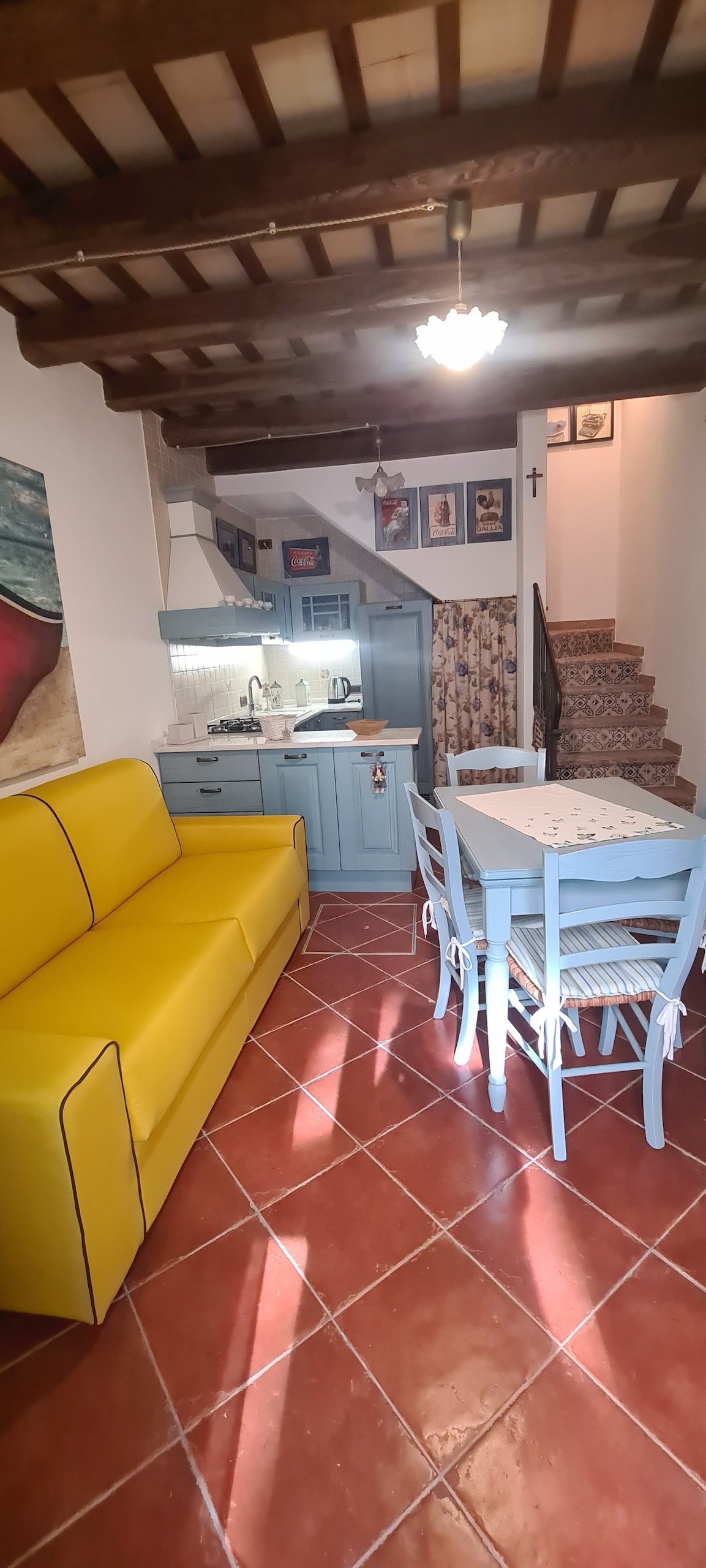A compact living space features a bright yellow sofa, a light blue dining table surrounded by chairs, and a kitchen area visible in the background. Warm wooden beams adorn the ceiling, and patterned tiles lead to a staircase that ascends to the upper level.