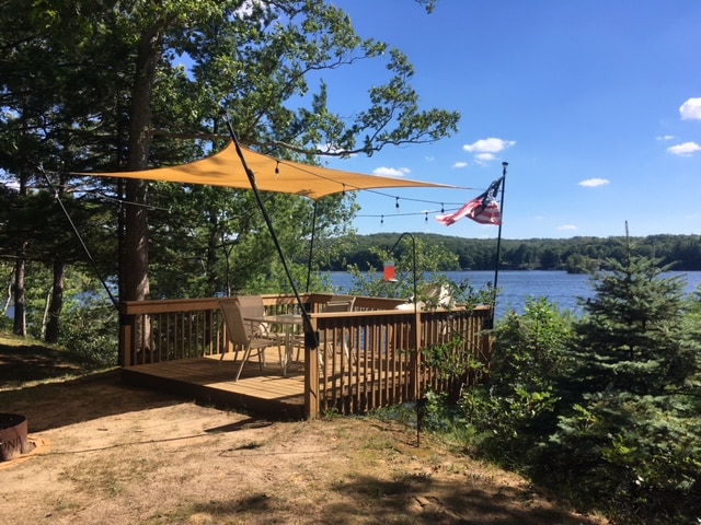 A wooden deck overlooking Croton Pond is equipped with seating arrangements and a shade canopy. An American flag is displayed alongside trees, enhancing the natural surroundings. The tranquil water view is framed by greenery, creating a peaceful outdoor space for relaxation.