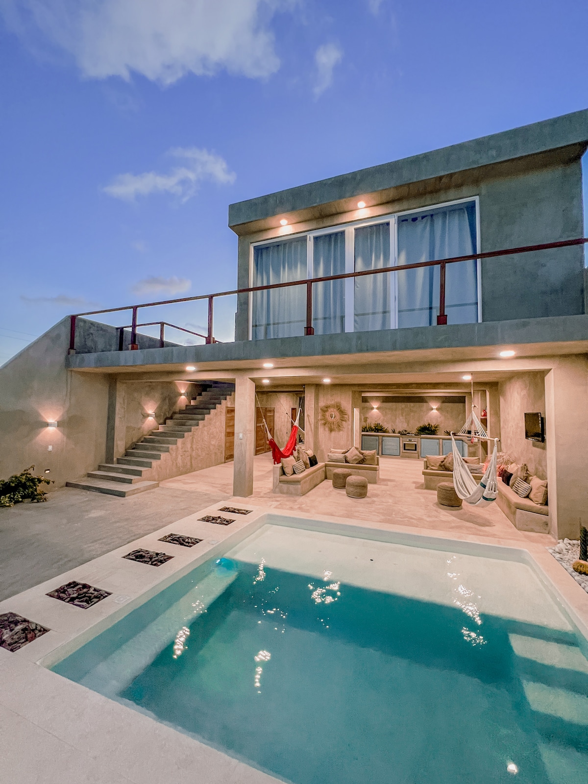 A modern villa with an inviting outdoor area is highlighted by a private pool, surrounded by light stone tiles. Hammocks are draped for relaxation beneath a covered patio, featuring casual seating. Expansive glass doors offer a view into the well-lit interior space at dusk.