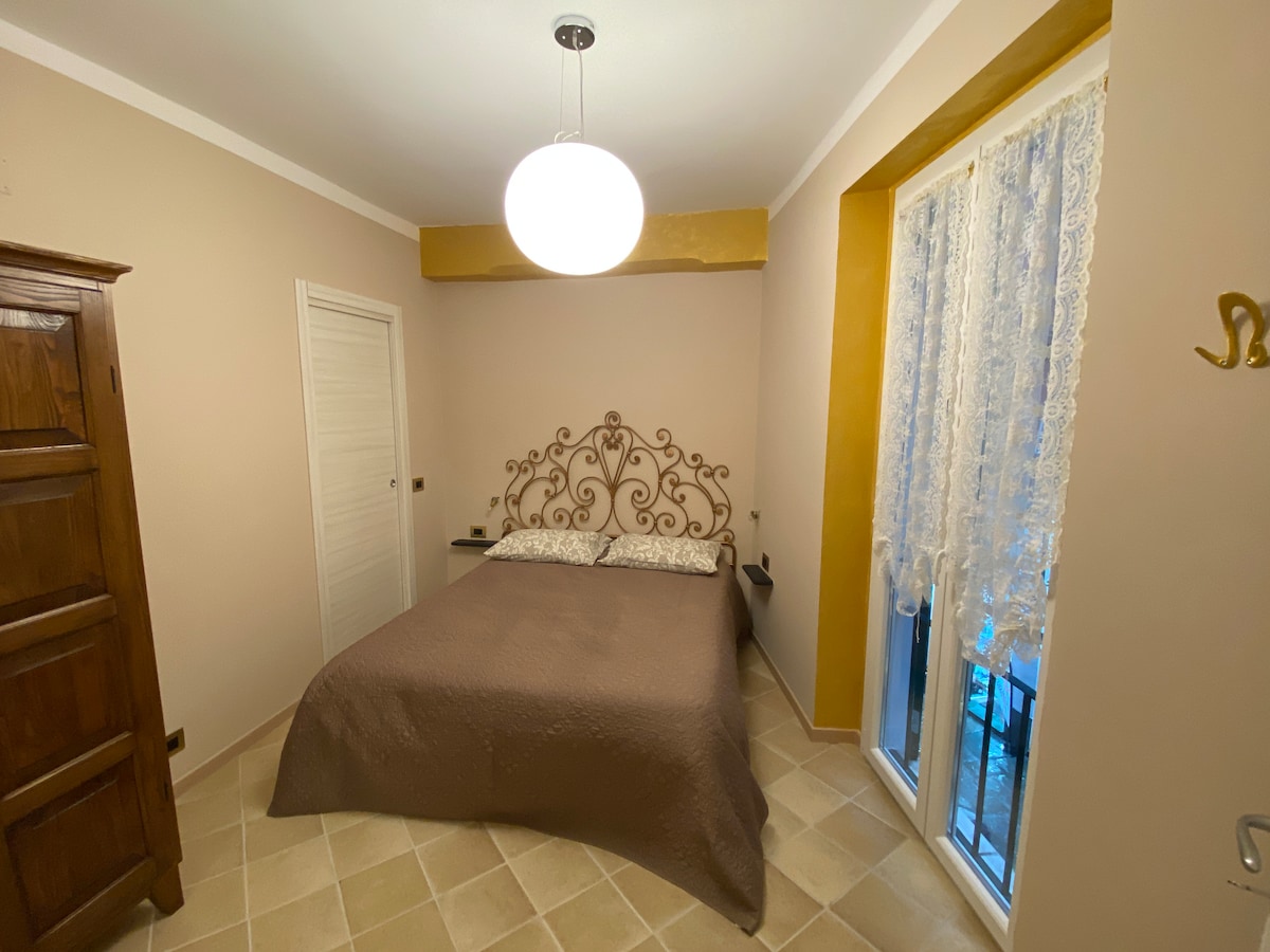 A comfortable bedroom is depicted, featuring a spacious bed with a decorative headboard. The walls are in soft beige tones, and a large window with lace curtains allows natural light to fill the room. An elegant wooden armoire is positioned nearby.