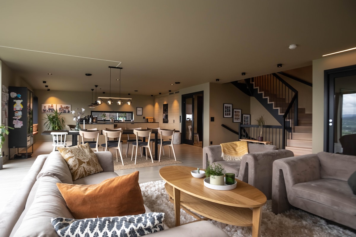 An open living area features a comfortable seating arrangement with light-colored sofas and decorative cushions. A wooden coffee table sits at the center, surrounded by a soft rug. The modern kitchen and dining area are visible in the background, with sleek countertops and inviting lighting.