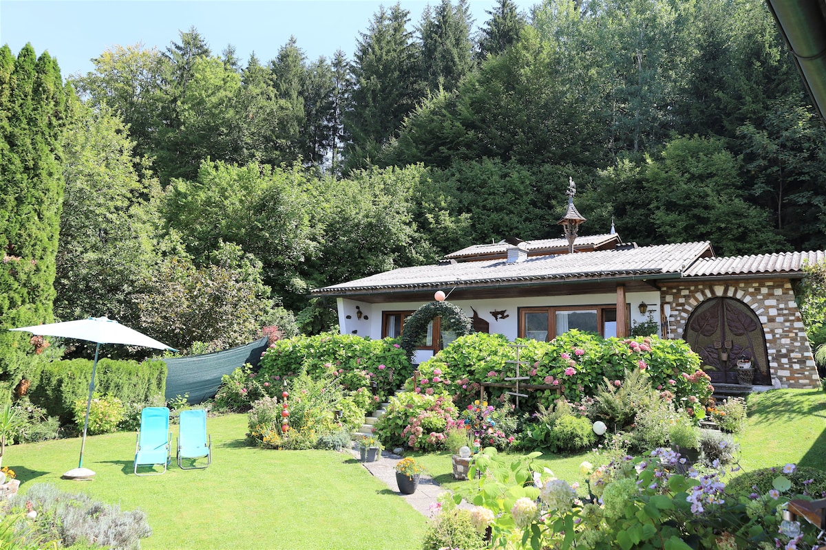 A charming bungalow is nestled in a lush garden, surrounded by vibrant flowers and greenery. Two lounge chairs are positioned on the lawn, while a sun umbrella provides shade. The backdrop features trees and a well-maintained pathway leading to the entrance of the bungalow.