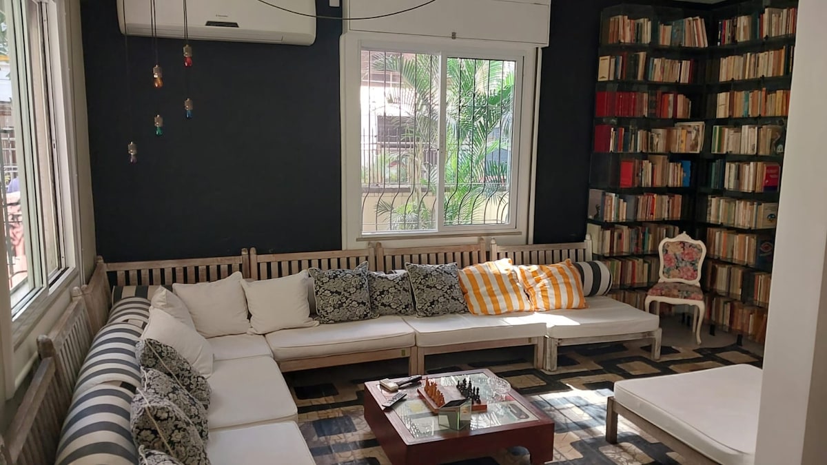 A cozy living area features a spacious sectional sofa adorned with various patterned cushions. A wooden coffee table is centered on a layered rug, with a chess set arranged for play. A wall of bookshelves frames the space, and large windows allow natural light to enter.