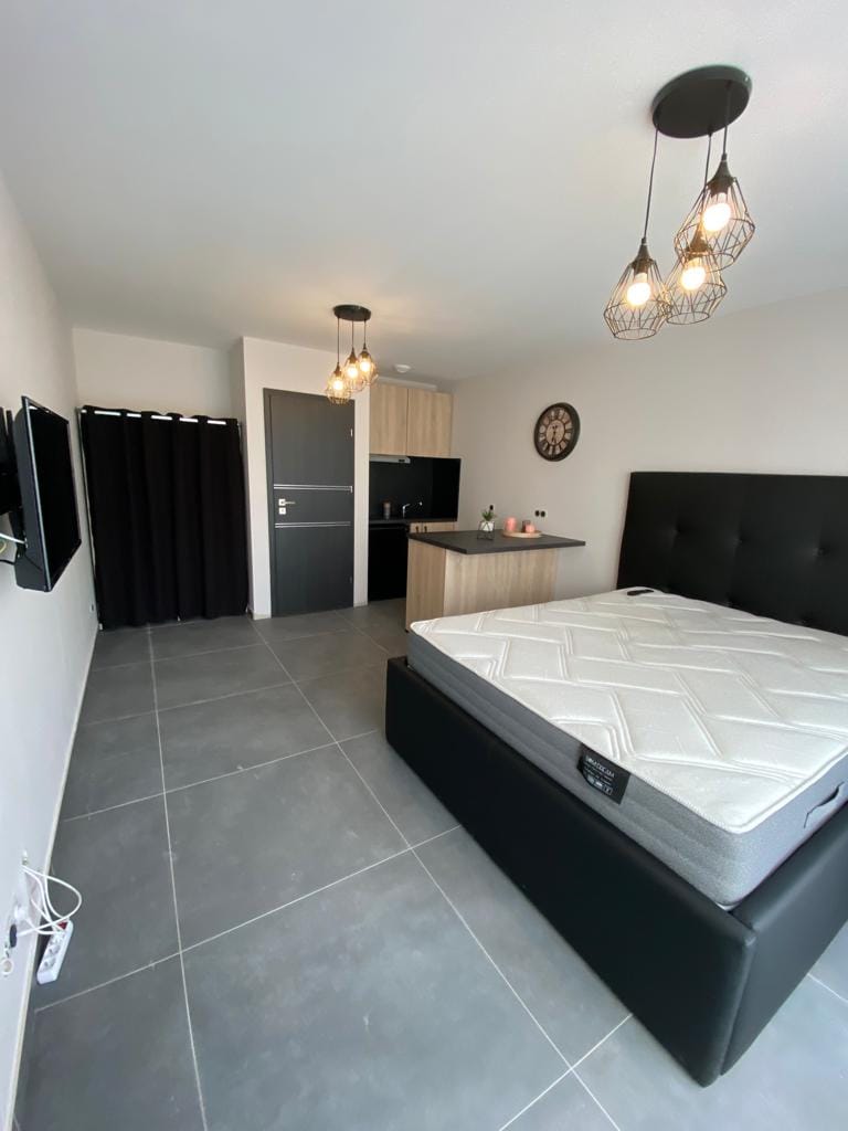 A spacious studio features a dark-colored bed frame positioned against a wall, with an unmade mattress. Soft lighting is provided by pendant lights above, while a kitchenette is visible in the background. A modern television is mounted on the wall, and a partial room divider offers privacy.