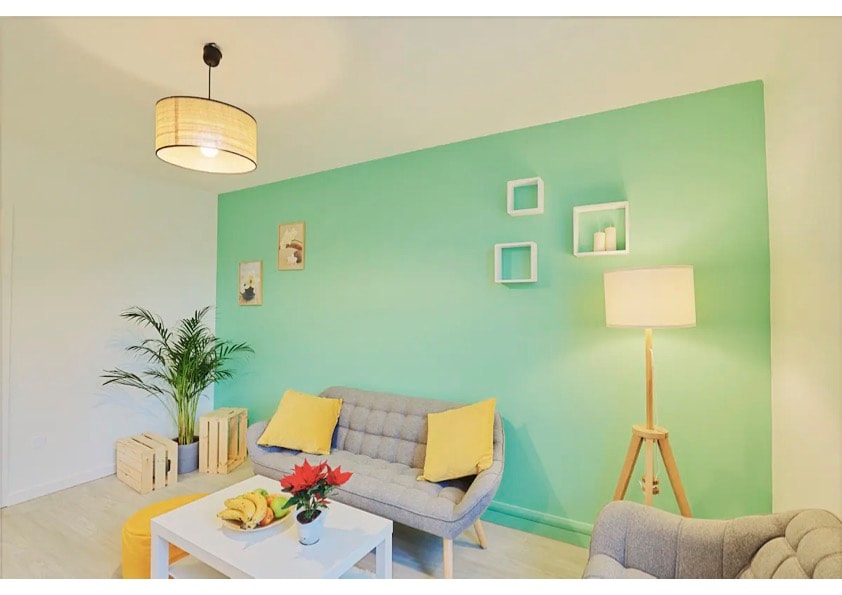 A bright living area features a green accent wall, complemented by light-colored furniture. A gray sofa and a yellow-accented chair are positioned around a white coffee table. Decorative wall shelves display simple decor. A floor lamp adds soft light, while a plant brings a touch of nature.