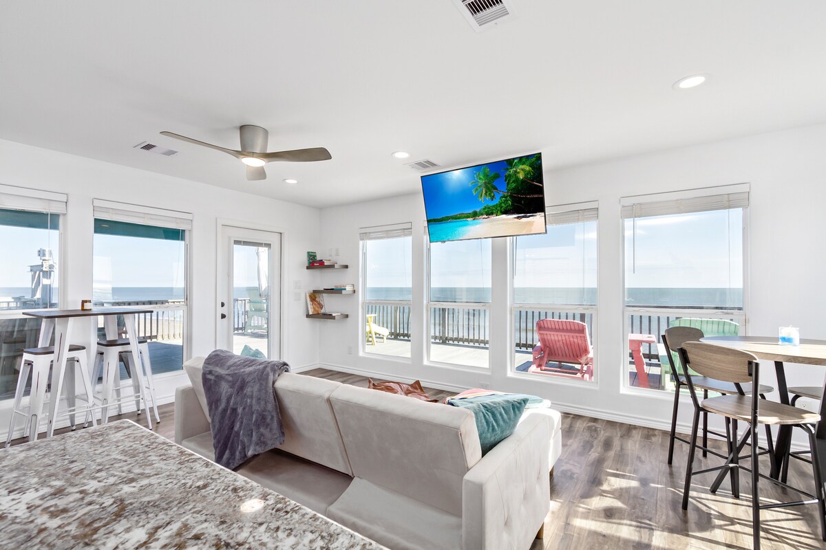 A bright living space features large windows with ocean views and a spacious sectional sofa. A mounted flat-screen TV is visible, and various seating options, including barstools and dining chairs, are arranged around a wooden table. Natural light enhances the open atmosphere.