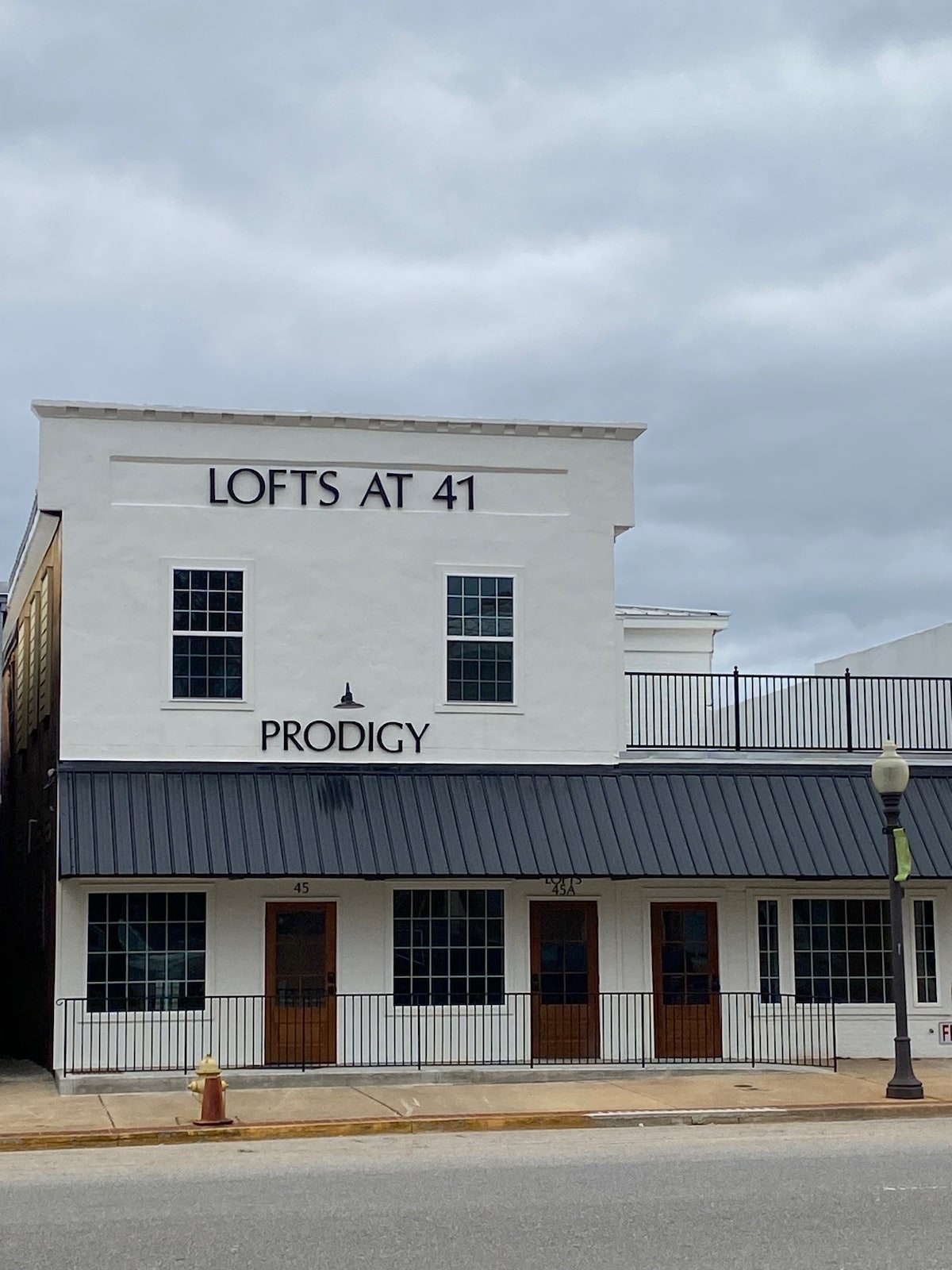 The building showcases a modern two-story structure with a white facade and a contrasting dark metal roof. Large windows line the front, allowing ample natural light. The name 'LOFTS AT 41' is prominently displayed above the entrance, which features wooden doors.