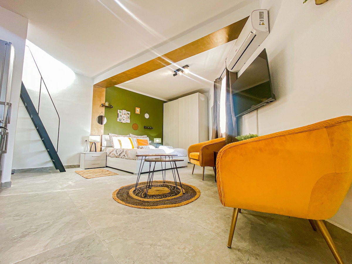 A modern studio apartment is illuminated by natural light. A comfortable king size bed is visible, alongside a cozy seating area featuring two bright yellow chairs. A flat screen TV is mounted on the wall, and a decorative rug is placed in the center of the room.