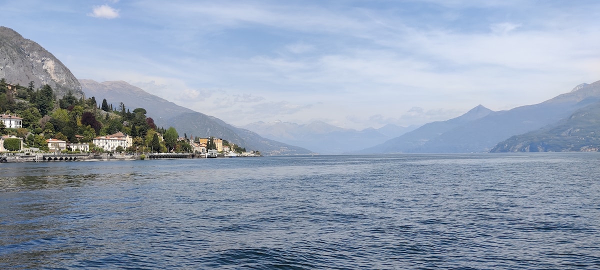 A serene view of Lake Como is captured, showcasing gentle waves reflecting the sky. The shoreline features a blend of lush greenery and charming buildings. Mountains rise in the background, creating a natural frame for the tranquil waters.