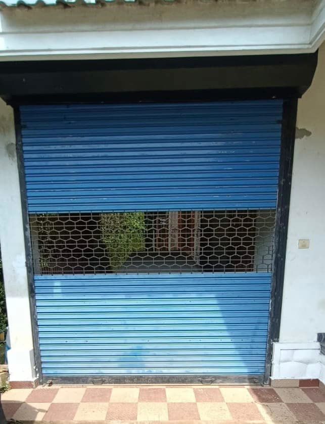 A blue roll-up security shutter is displayed, partially open to reveal a view of the interior space. The shutter's metal slats are arranged horizontally, and a grid pattern is seen through the openings. The ground features a tiled pattern in neutral colors.