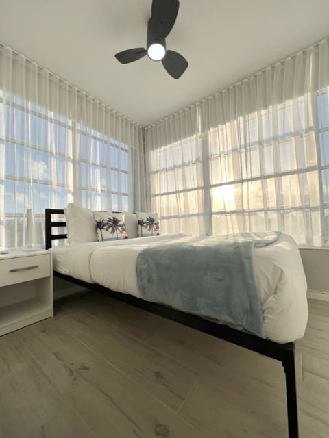A well-lit bedroom features large windows adorned with sheer curtains, allowing natural light to filter through. A spacious bed is dressed with neutral linens and decorative pillows. A bedside table is present, and a ceiling fan is positioned above for added comfort.