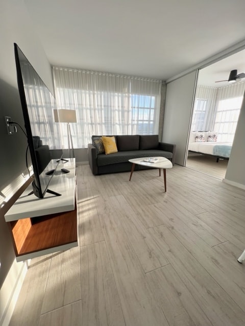 A modern living area is shown, featuring a dark sofa with colorful accent pillows and a light wooden coffee table. A wall-mounted TV is positioned above a sleek cabinet. Large windows with sheer curtains allow ample natural light to brighten the space.
