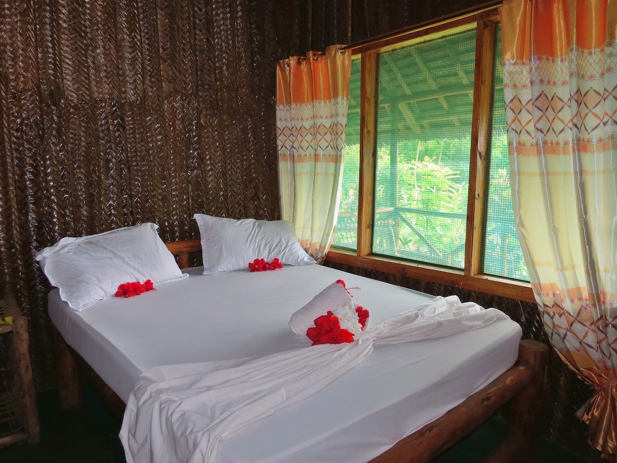 A spacious bed adorned with white linens and decorative towels lies in a rustic room. Large windows with light curtains allow natural light to flow in, offering views of the lush surroundings. The walls are covered with natural materials, creating a warm ambiance.