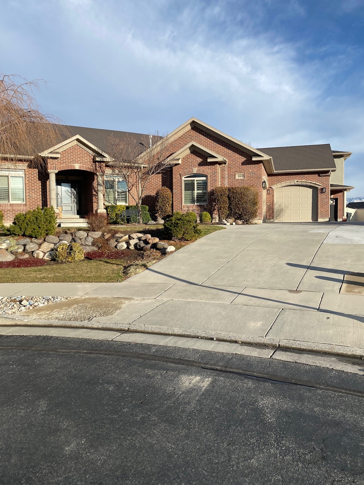 A welcoming exterior view of a well-maintained brick home is displayed, featuring a landscaped front yard with shrubs and rocks. The driveway slopes gently, leading to a private garage on the right, with the entrance steps visible, framed by greenery.