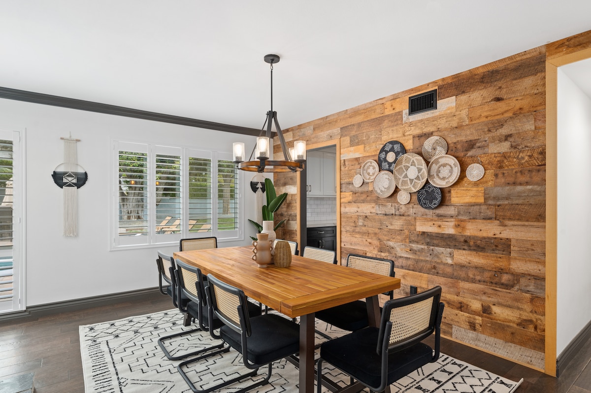 A dining area features a long, wooden table surrounded by six chairs. A decorative wall showcases an arrangement of plates against a natural wood backdrop. Light streams in through glass doors, providing views of the outdoor pool area.