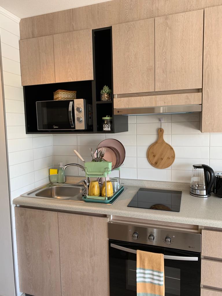 A compact kitchen area features modern, wood-toned cabinetry and a stainless steel sink. An electric stove is adjacent to a microwave. A cutting board and decorative items are visible, alongside a kettle and coffee maker, creating a functional cooking space.