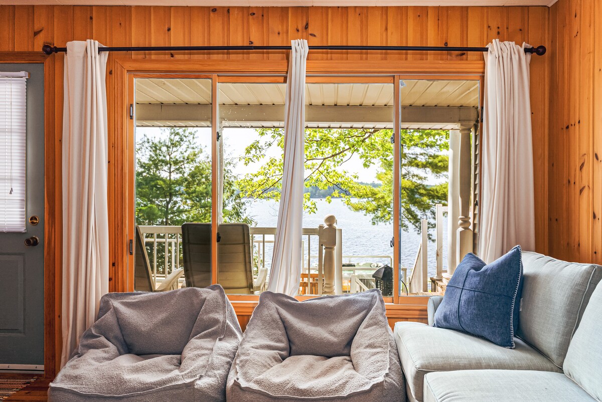 A cozy seating area is presented with soft bean bag chairs positioned in front of large glass doors. The doors open to a scenic view of Pine Lake, framed by natural wood walls and soft white drapes, inviting natural light into the space.