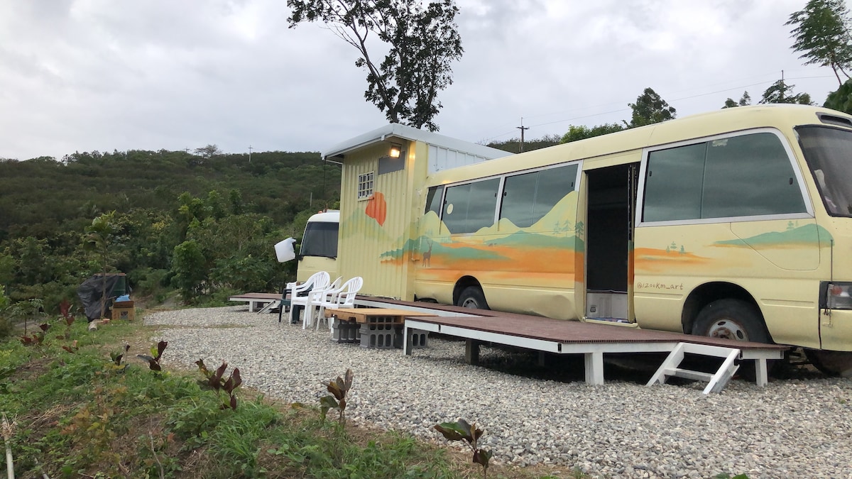 A vintage bus with a colorful mural is positioned on a gravel area. Nearby, two white chairs sit on a wooden deck, surrounded by lush greenery. The landscape features gentle hills and a cloudy sky, creating a calm atmosphere in the outdoor setting.