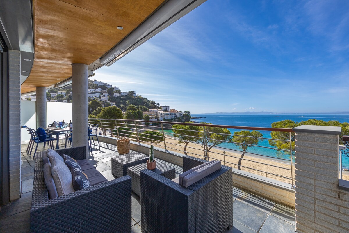 A spacious terrace showcases comfortable seating arrangements and a dining area, overlooking the sandy beach and coastal scenery. The clear blue sky enhances the inviting outdoor space, while trees provide a natural backdrop, creating a serene environment for relaxation.