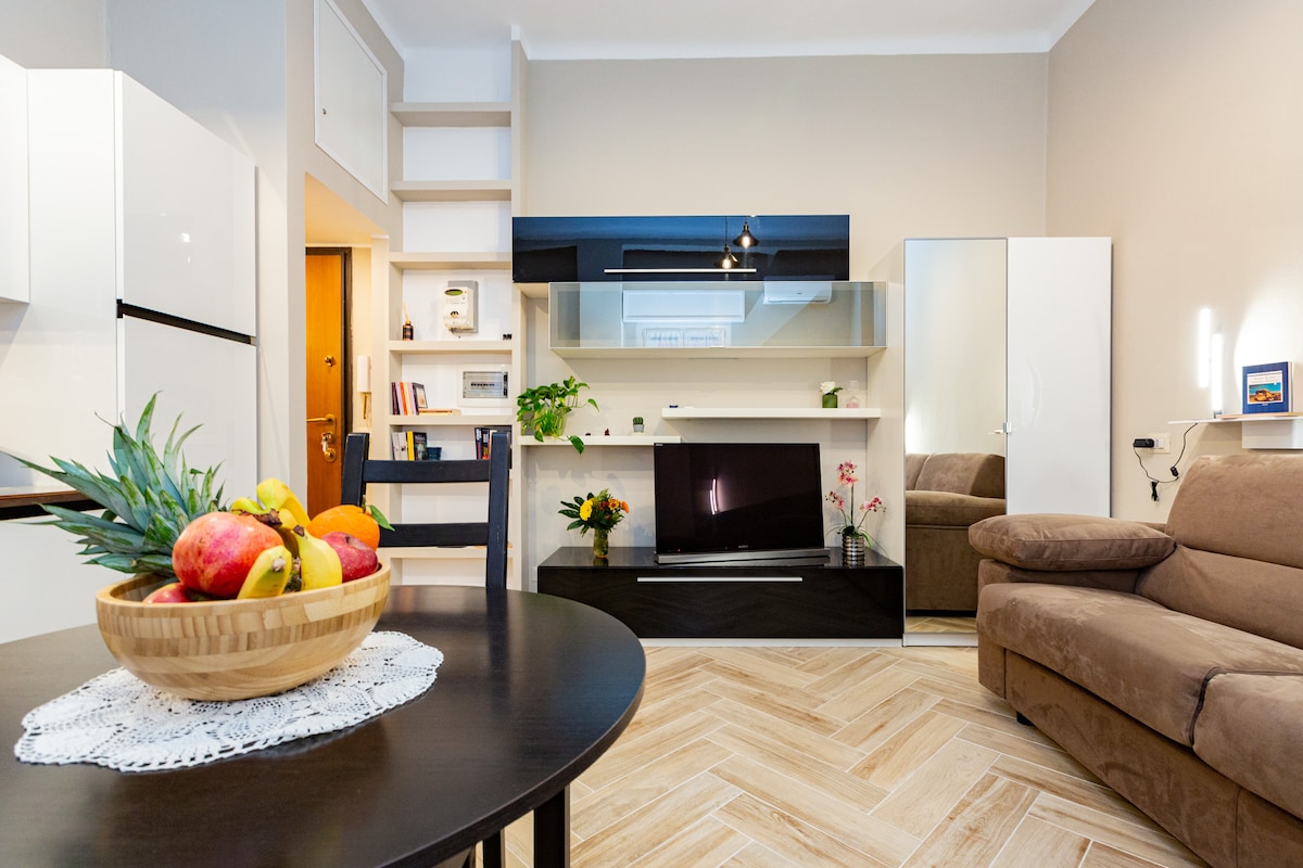 A modern living area features a comfortable brown sofa alongside a sleek, low-profile television stand. A dining table is adorned with a bowl of fresh fruit, while decorative plants add a touch of greenery. Natural light filters through the space, enhancing its welcoming ambiance.