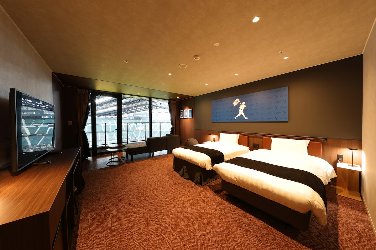 A spacious room is furnished with two semi-double beds and a flat-screen television. Large windows provide an unobstructed view of the Escon Field. Artwork adorns the walls, adding character to the comfortable setting.