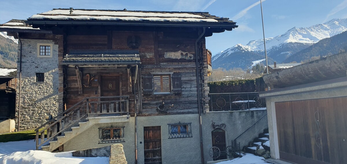 A traditional chalet with a wooden façade is situated against a mountainous backdrop. Snow covers a small lawn area, while stairs lead to the main entrance. A balcony above the entrance features rustic wooden railings, and windows display local architectural elements.