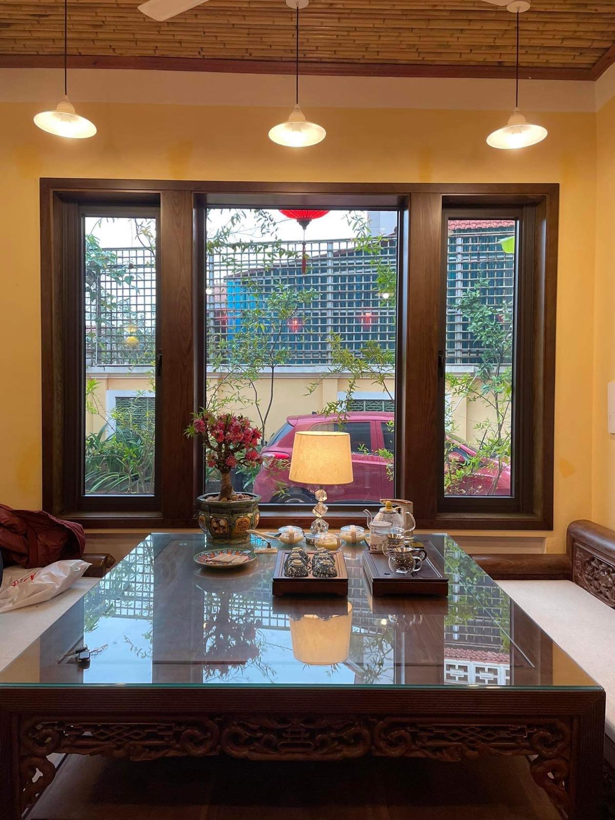 Large windows frame views of an outdoor garden, allowing natural light to fill the space. A wooden table is set with decorative items, including a lamp and a vase of flowers, contributing to a bright and inviting ambiance.
