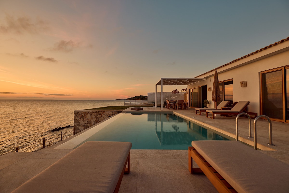 An outdoor area features a spacious swimming pool with steps leading in, set against a sunset-lit sea. Two sunbeds are arranged nearby, and a shaded dining space is visible under a pergola, enhancing the serene atmosphere of the villa's private surroundings.