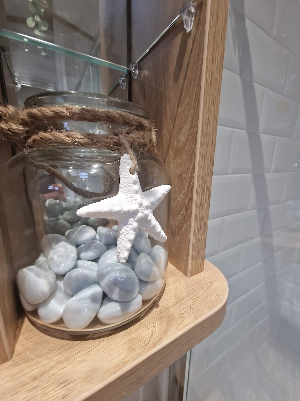 A glass jar filled with smooth stones sits on a wooden shelf. The jar is adorned with a piece of twine and a white starfish hanging from it, adding a decorative touch to the neutral-toned surroundings.