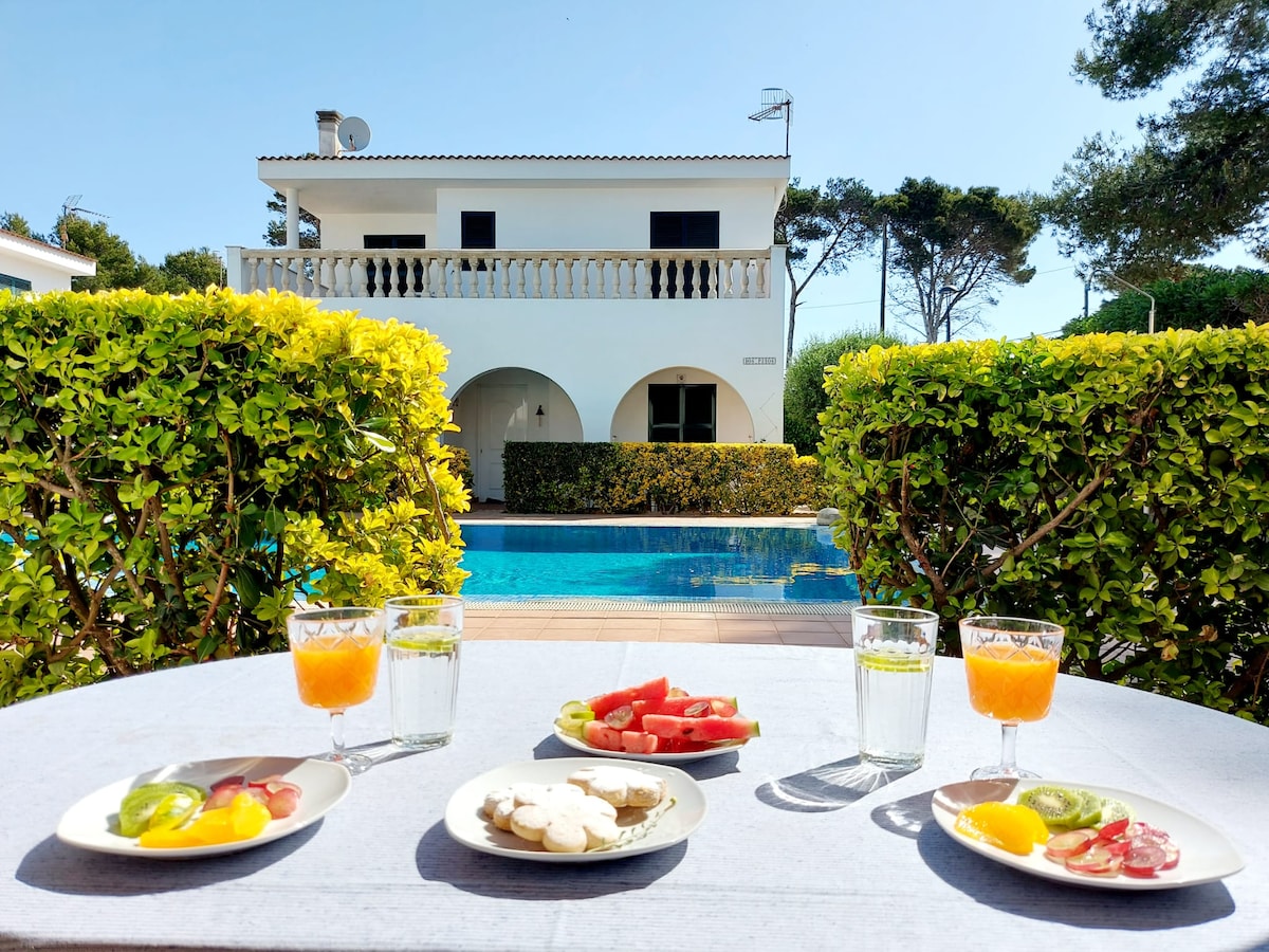 A table is set outdoors, featuring plates of fresh fruit and two glasses of orange juice alongside clear water. The background shows a turquoise pool and a two-story villa framed by lush greenery and blooming bushes under a clear blue sky.