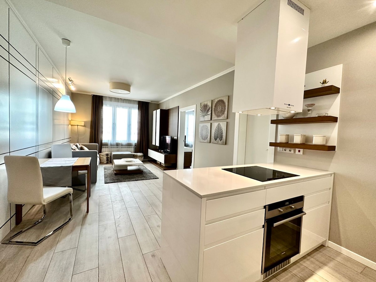 A modern kitchen area is showcased with sleek white cabinetry and an island featuring a built-in cooktop. The space is open to a living area, where a comfortable sofa and chic coffee table are positioned. Large windows allow natural light to fill the room.