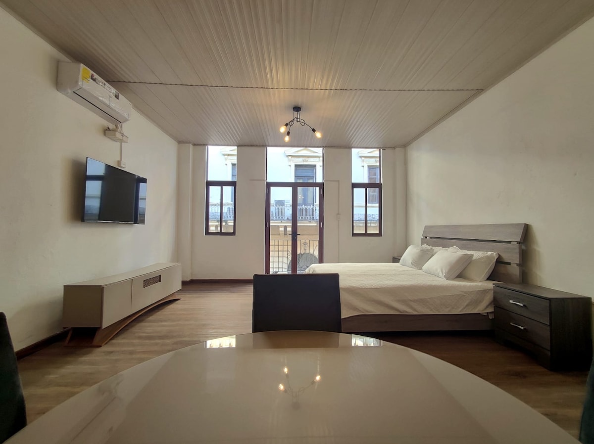 The studio features a spacious layout with large windows allowing ample natural light. A comfortable bed is placed near the windows, and a smart TV is mounted on the wall. A dining table for four is also visible in the foreground.