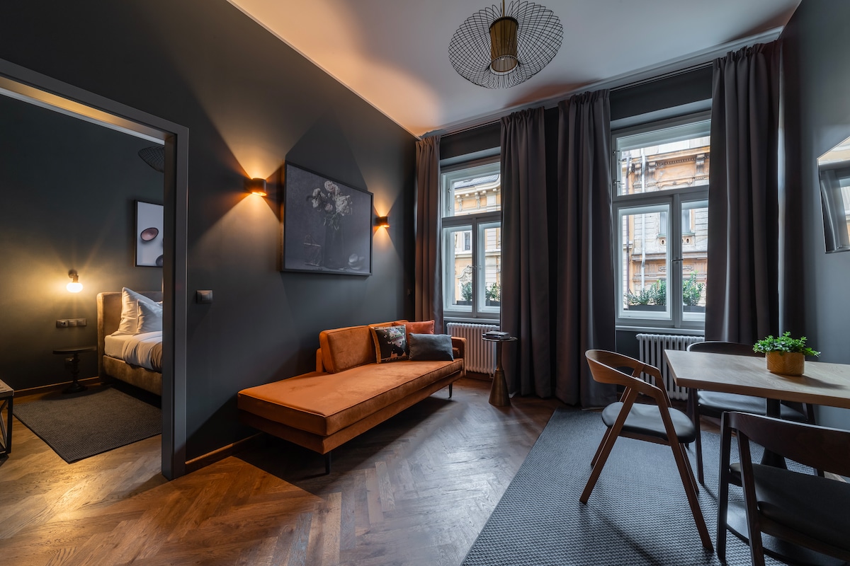 A separate living area is illuminated by warm lighting, featuring a stylish orange sofa and a wooden dining table with chairs. Large windows covered by dark curtains allow natural light to enhance the room's modern decor. A cozy bedroom is visible through the open doorway.
