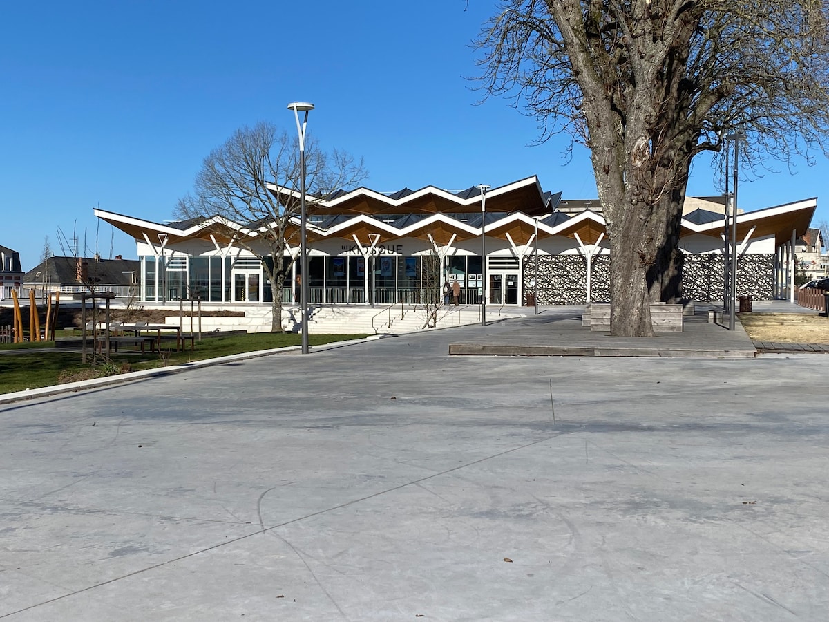 A modern building features a distinctive zigzag roof design, surrounded by landscaped green space. The façade is composed of large glass windows, offering a bright, open atmosphere. Nearby, outdoor seating areas and trees complement the spacious plaza in front.