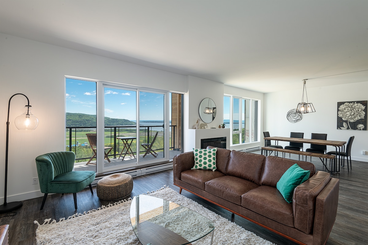 A spacious living area features a large brown leather sofa, complemented by a green accent pillow. A coffee table sits on a textured rug. Floor-to-ceiling windows offer a view of the surrounding landscape, while a dining area with a table for six is visible in the background.