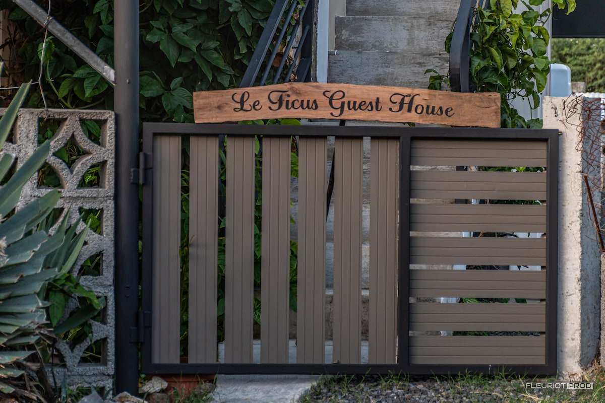 A wooden sign labeled 'Le Ficus Guest House' is displayed above a modern gate. The gate features horizontal slats and is complemented by greenery surrounding the entrance, providing a welcoming access point to the property.