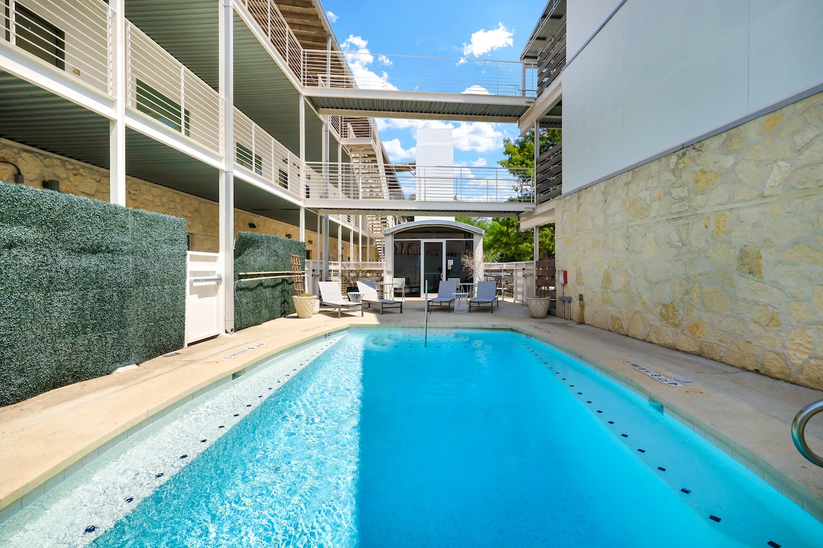 A tranquil pool area is surrounded by a stone patio featuring lounge chairs. Sunlight reflects off the clear water, highlighting the inviting pool design. A landscaped privacy wall is present, complementing the overall aesthetic of the outdoor space.