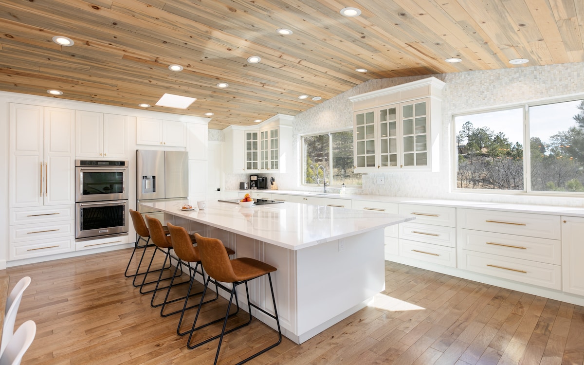 A spacious kitchen features a large island with seating for five. White cabinetry and countertops are complemented by warm wooden ceiling beams. Natural light fills the space through large windows, showcasing views of the surrounding landscape.