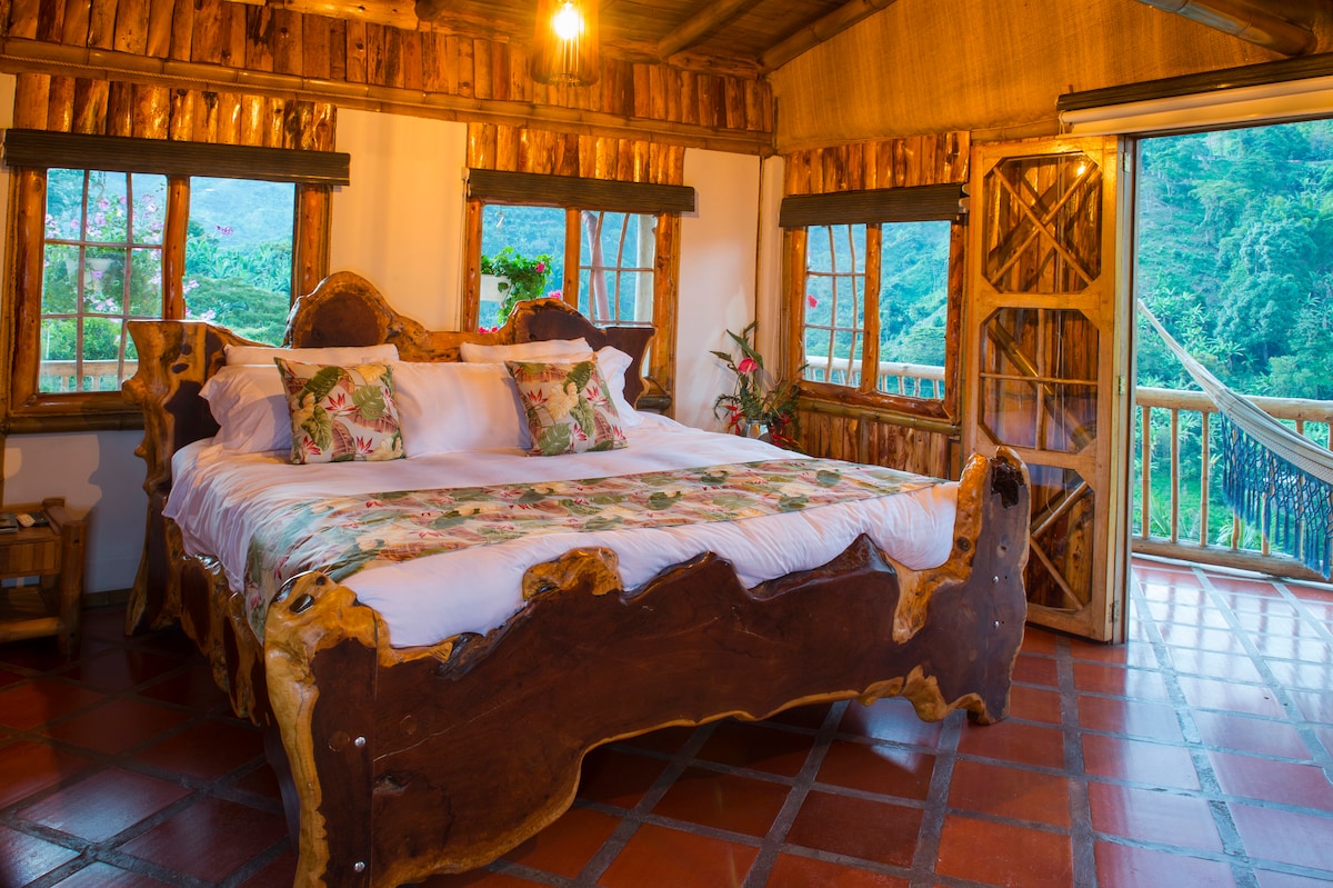 A spacious bedroom is furnished with a handcrafted wooden bed adorned with floral bedding. Large windows frame picturesque views of the mountains, allowing natural light to brighten the room. A small seating area is present, complemented by rustic wooden accents.
