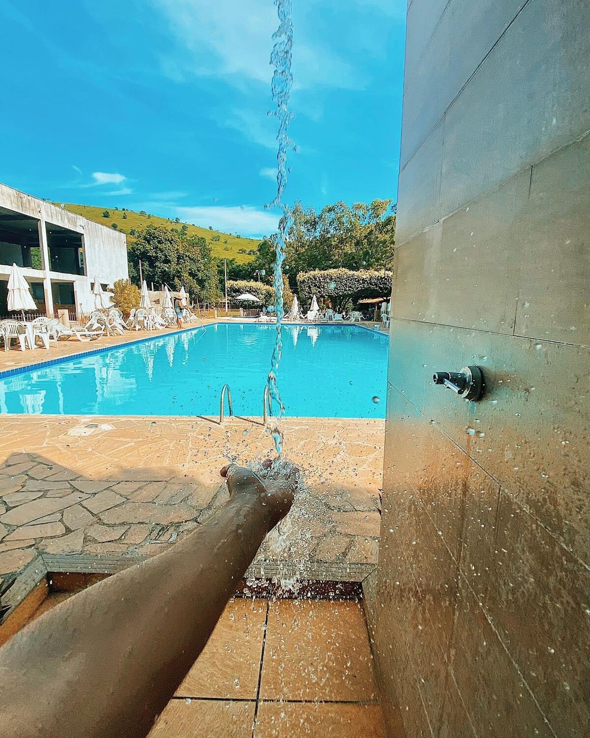 A hand is positioned under a cascading water source, with a clear blue pool visible in the background surrounded by lounge chairs and umbrellas. Lush greenery and a sunny sky are seen beyond the pool area, creating a serene atmosphere.