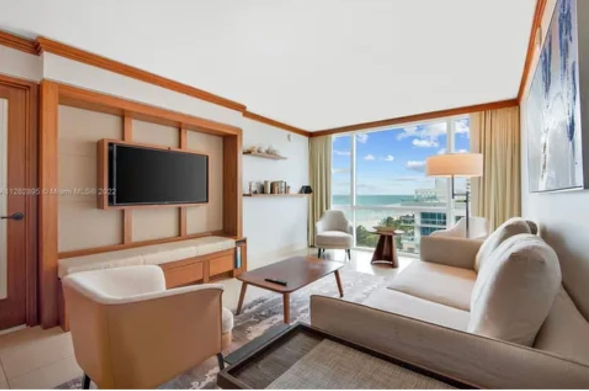 The living area features a comfortable seating arrangement including a light-colored sofa and armchairs around a wooden coffee table. A flat-screen television is mounted on the wall, and large windows offer an unobstructed view of the ocean and sky.