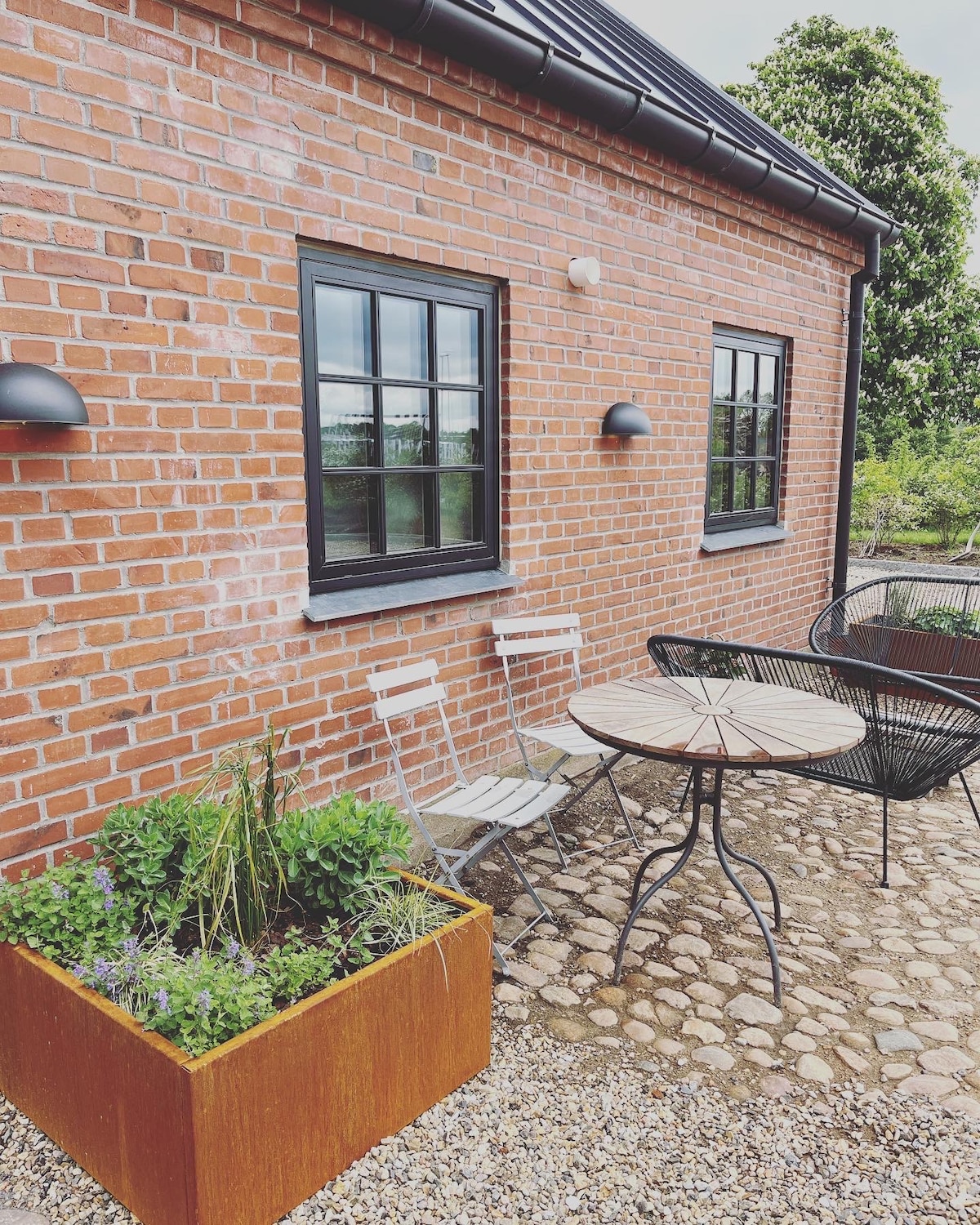 A charming outdoor seating area is visible, featuring a round table and two chairs against a backdrop of a brick wall. Lush green plants are arranged in a decorative planter, while the path is made of smooth cobblestones, creating a welcoming environment.