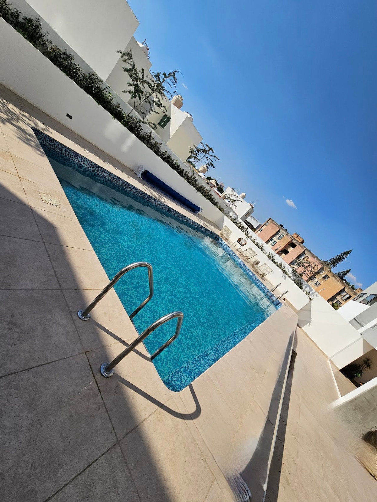 A serene swimming pool is set within a modern outdoor space. Clear blue water reflects the sky, while smooth beige tiles surround the pool. Lounge chairs are positioned along the edge, providing relaxation areas, with green trees and buildings visible in the background.