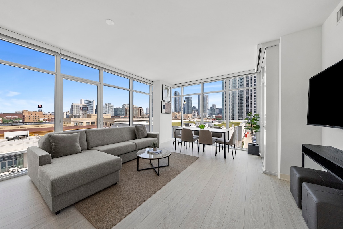 A modern living space is displayed with expansive windows offering a view of the Chicago skyline. A comfortable gray sectional sofa and a round coffee table are positioned on a light-colored rug. A dining area with a table and chairs is visible in the background.