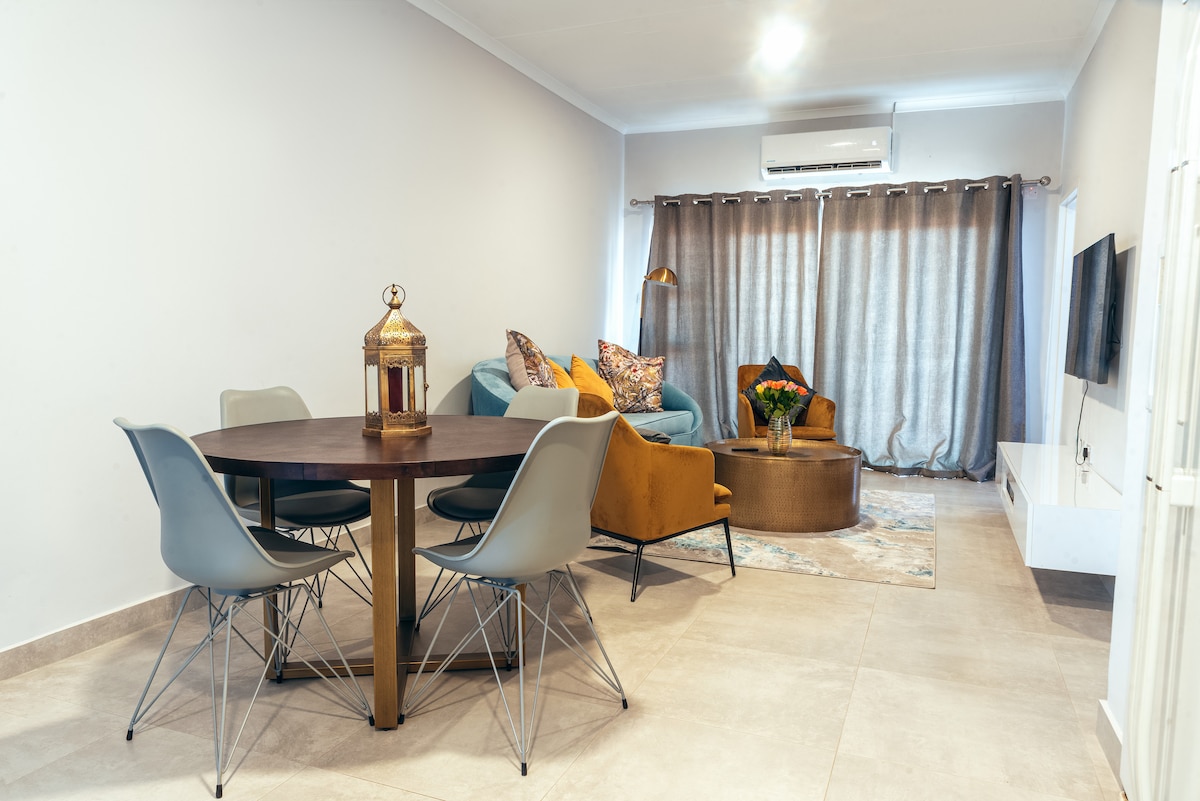 A modern living area features a round dining table with four chairs, complemented by a cozy seating arrangement. An armchair and sofa in shades of blue and gold are accented with colorful cushions. Natural light enters through large, draped windows, illuminating the neutral-toned walls.
