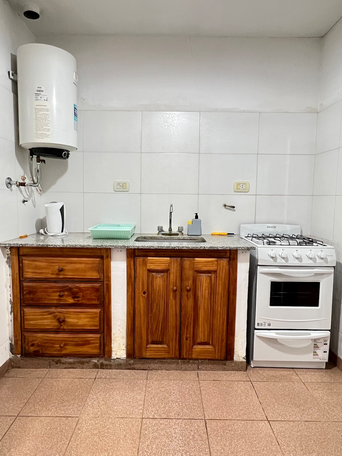A functional kitchen space features wooden cabinets and a countertop with a sink. A white gas stove is positioned beside the cabinets. Natural light illuminates the area, enhancing the simplicity of the tile flooring and the overall clean design.