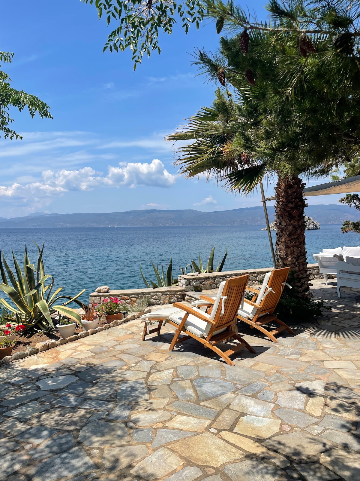 Two wooden loungers are positioned on a stone patio overlooking the sea. Lush greenery and vibrant flowers surround the area, while distant mountains can be seen across the water under a partly cloudy sky.
